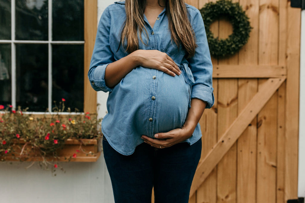 heavily pregnant woman wearing blue denim shirt and black trousers standing in front of a wooden gate